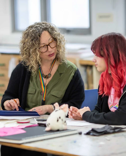 Lecturer with curly hair and glasses discussing an art project with a student who has red hair, in a classroom setting. Lecturer with curly hair and glasses discussing an art project with a student who has red hair, in a classroom setting.